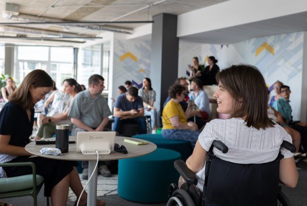 Kerekesszékes nő mosolyog egy modern irodai rendezvényen, ahol a háttérben többen ülnek és figyelnek egy előadást vagy workshopot.
