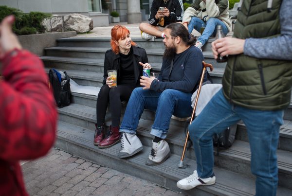 Kültéri lépcsőn ülő, vörös hajú nő és egy botra támaszkodó szakállas férfi kötetlen párbeszéde egy rendezvényen.