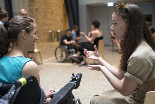 Elektromos kerekesszékben ülő, mosolygó nő és a vele szemben ülő, lelkesen magyarázó fiatal nő közvetlen párbeszéde egy rendezvényen.